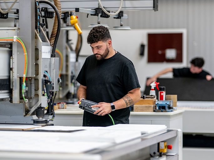 Ein junger Mensch arbeitet an einem Werktisch und steuert eine Maschine, die Kunststoffe präzise zuschneidet.
